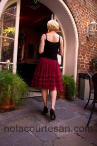 New Orleans Escort and Companion Annie Calhoun / Description: Back view of a woman with blonde hair and fair skin, wearing high heels, a skimpy black top, and a ruffled wine-colored skirt, walking through a French Quarter courtyard. 